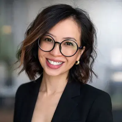 Professional headshot of a smiling person wearing round glasses and a black blazer against a blurred office background.